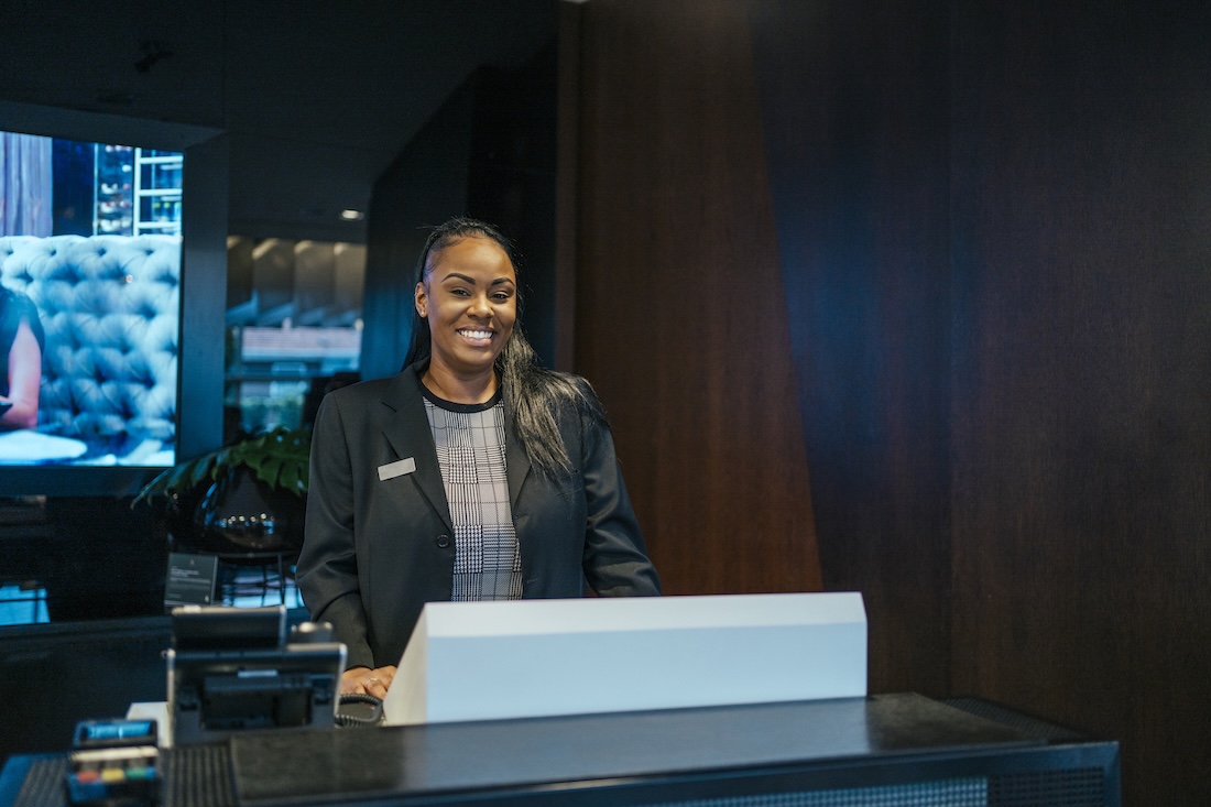 Smiling female hotel receptionist standing behind a front desk in a modern lobby.