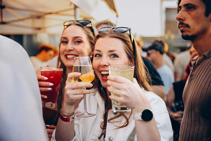 Zwei Frauen lachend mit Getränken in den Händen in einer Festival- oder Partyumgebung.