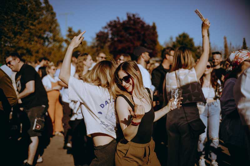 Zwei Frauen tanzen draußen in einer Menge bei sonnigem Wetter, eine trägt Sonnenbrille und hält ein Getränk.
