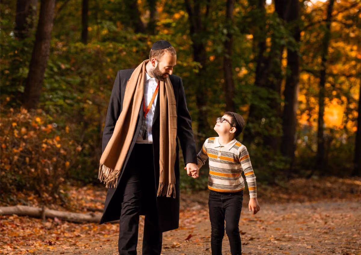 Man and boy wearing yarmulkes walking hand in hand on a forest path with autumn leaves.