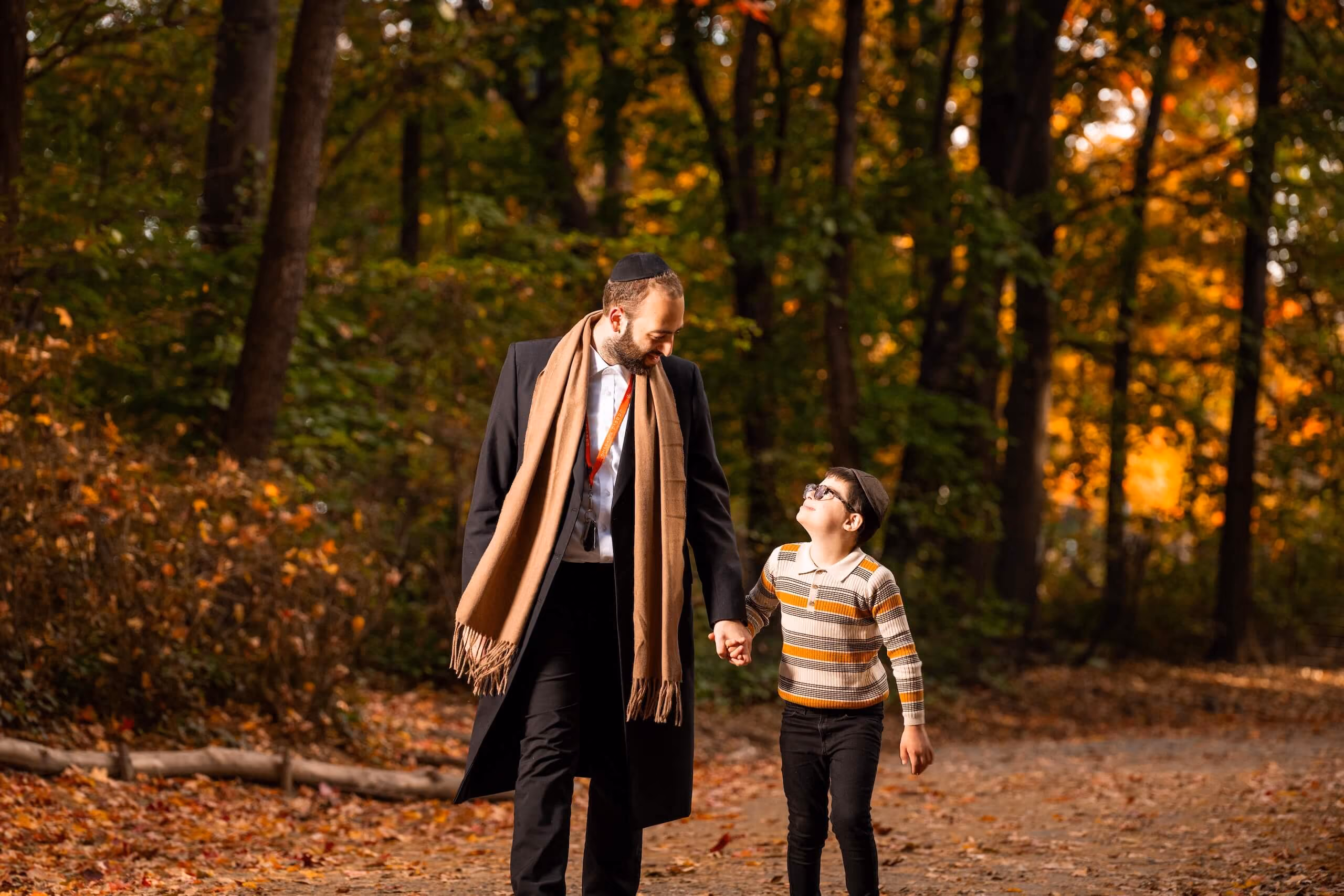 Man and boy wearing yarmulkes walking hand in hand on a forest path with autumn leaves.