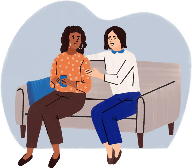 Two women sitting on a couch, one comforting the other who holds a cup.