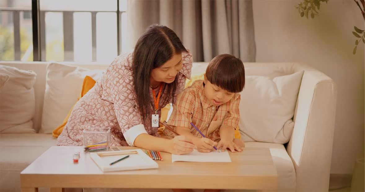 Woman helping young boy with drawing while sitting on a white couch at a wooden table.