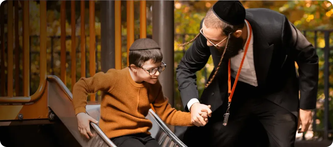 Man in black suit helping boy with glasses slide down playground slide outdoors.
