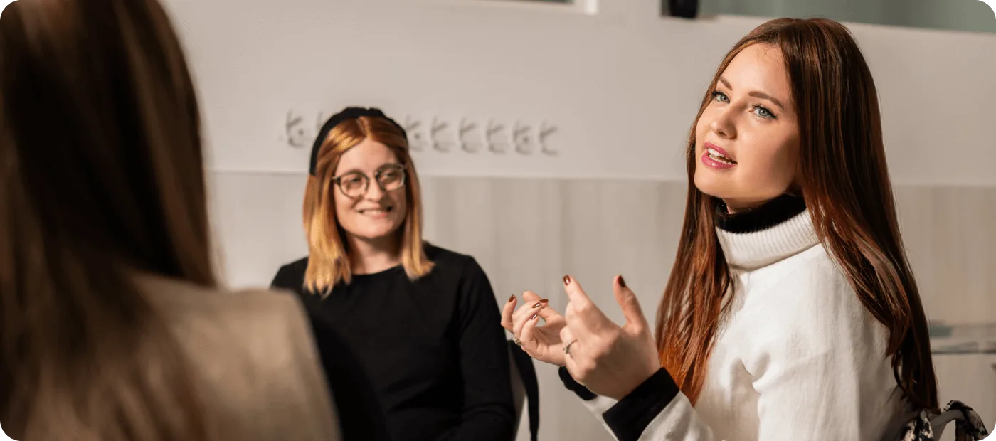 Two women in conversation, one speaking with hands raised, the other smiling and listening attentively.