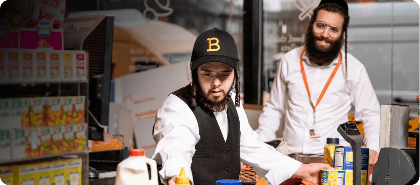 Man with black cap and vest stocking groceries at a store counter while another man watches behind.