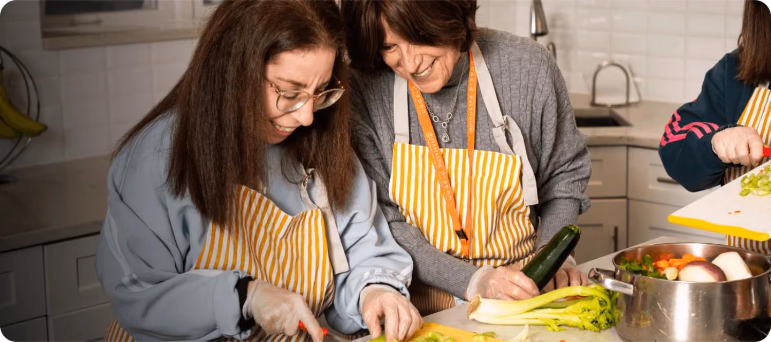 Two women wearing striped aprons cutting vegetables together in a kitchen.