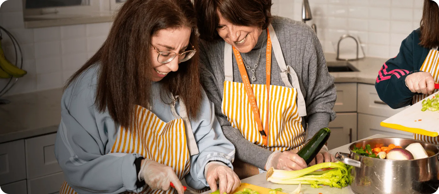 Two women wearing striped aprons cutting vegetables together in a kitchen.