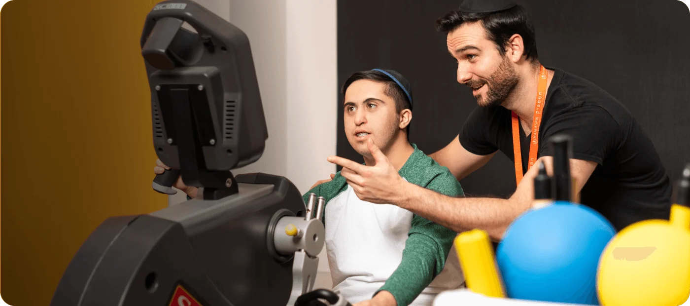 Man with kippah and a trainer using an exercise machine in a rehabilitation or habilitation clinic.