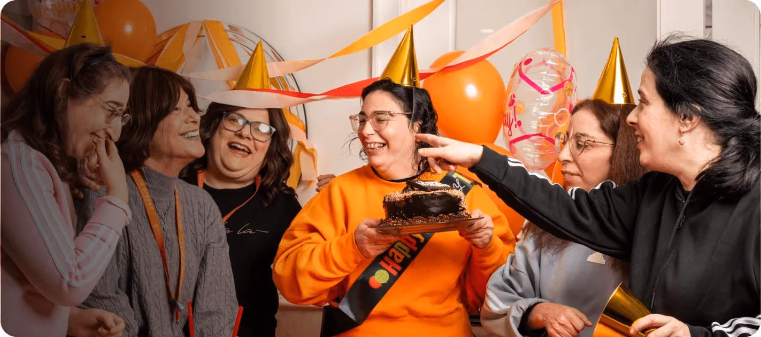 Group of women wearing party hats celebrating and holding a birthday cake and balloons indoors.