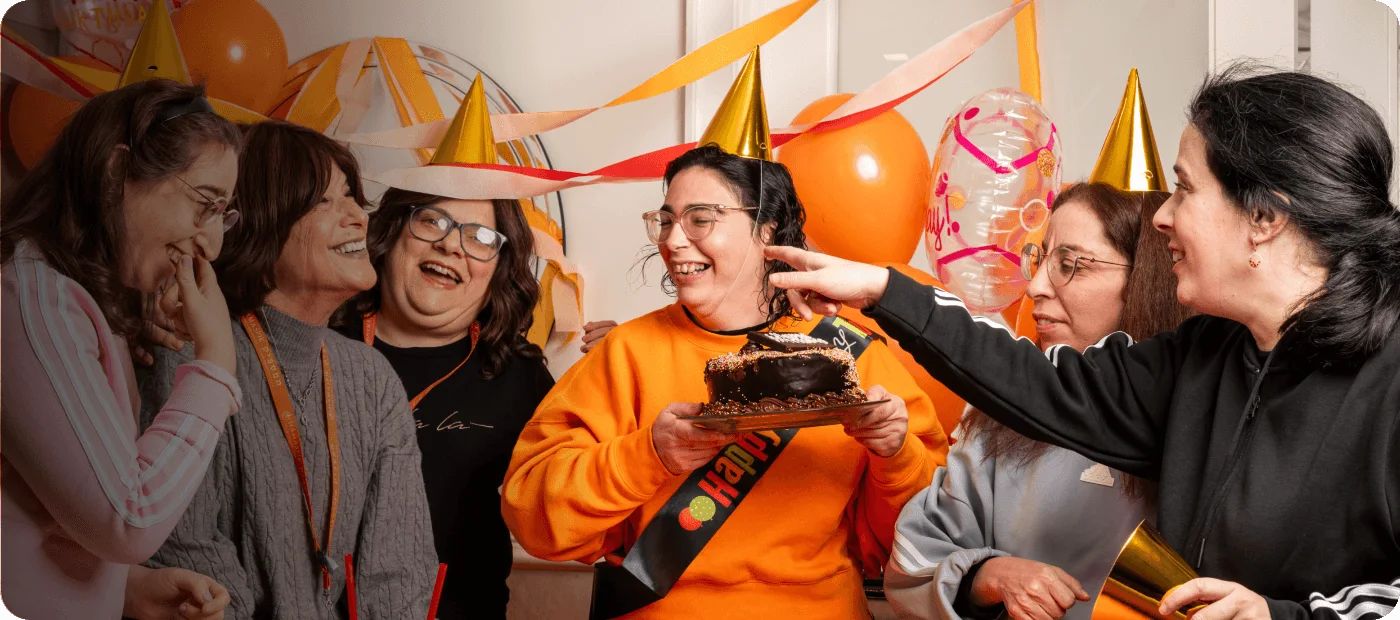 Group of women wearing party hats celebrating and holding a birthday cake and balloons indoors.