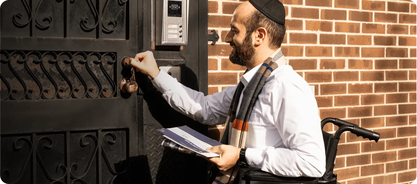 Bearded man in wheelchair knocking on black iron gate near brick wall, holding papers.