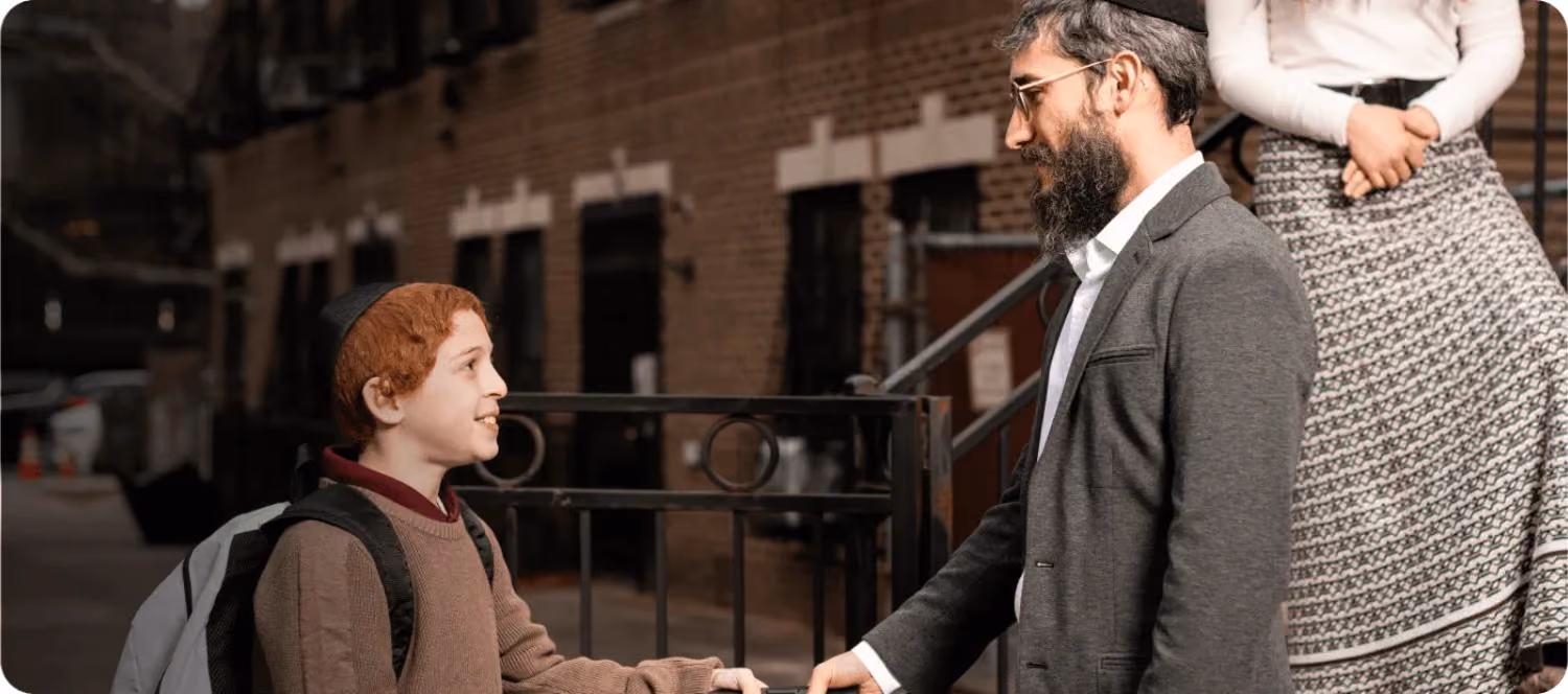 A boy with a backpack shakes hands with a bearded man wearing glasses and a kippah outdoors.