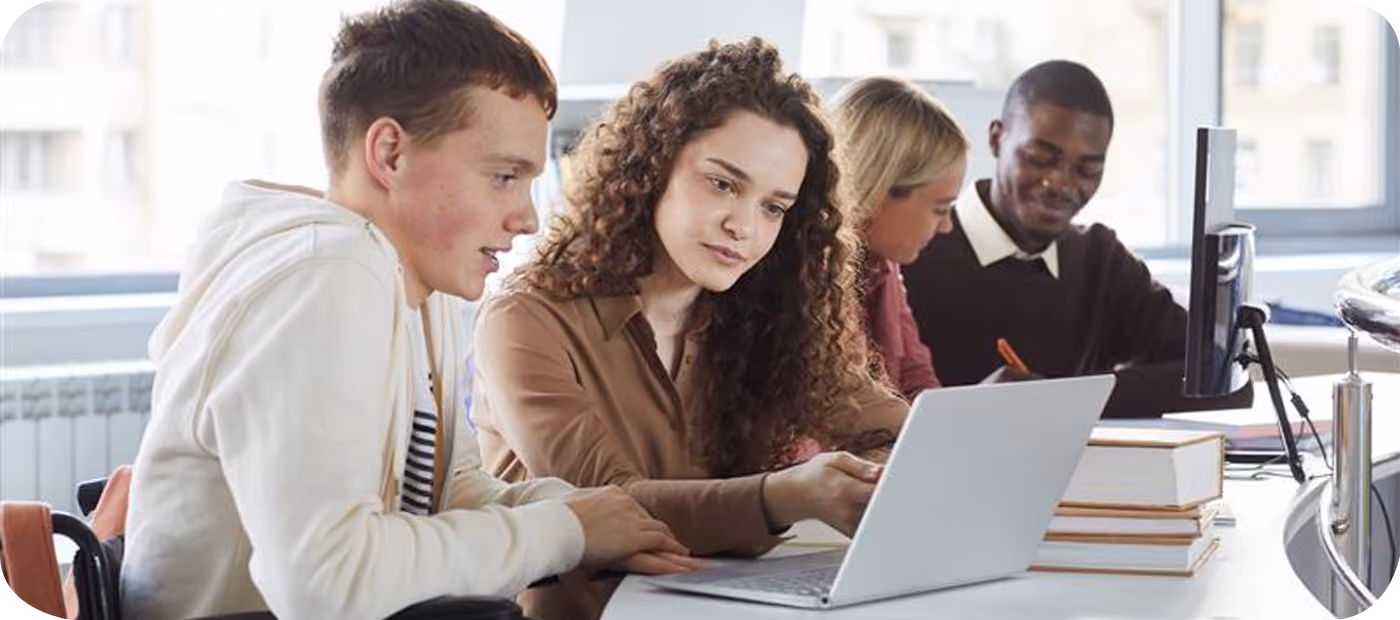 Two young adults closely collaborating on a laptop while two others work in the background in an office.