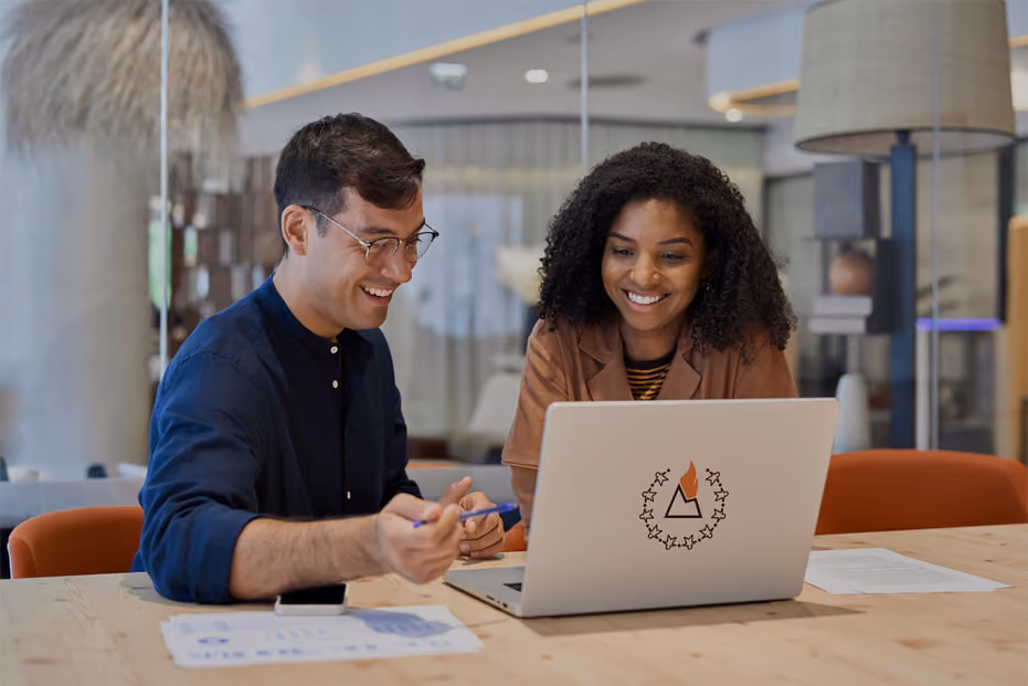 Two coworkers smiling and discussing work while looking at a laptop on a table in an office.
