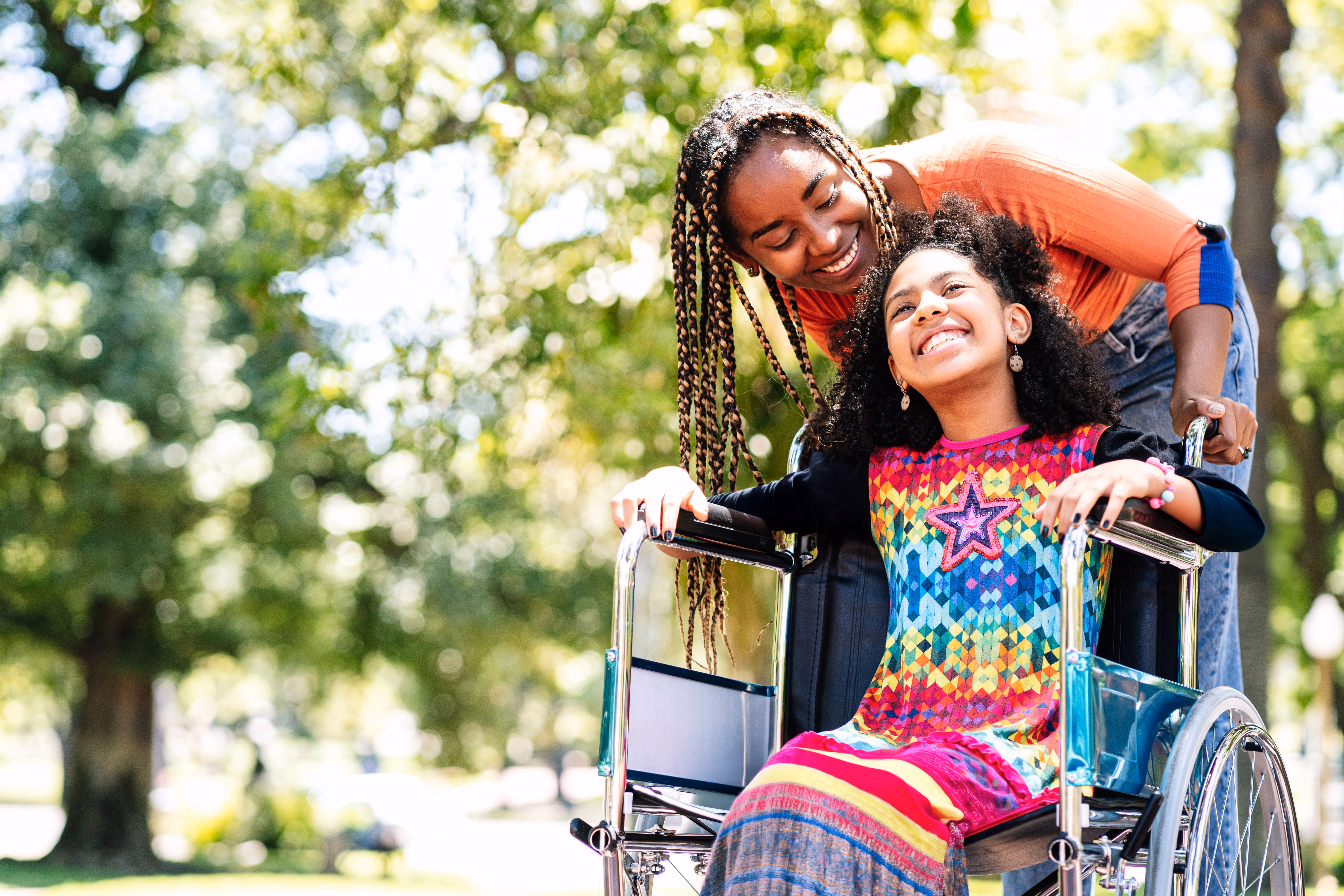 Smiling woman with braids pushes happy girl in colorful dress sitting in a wheelchair outdoors.