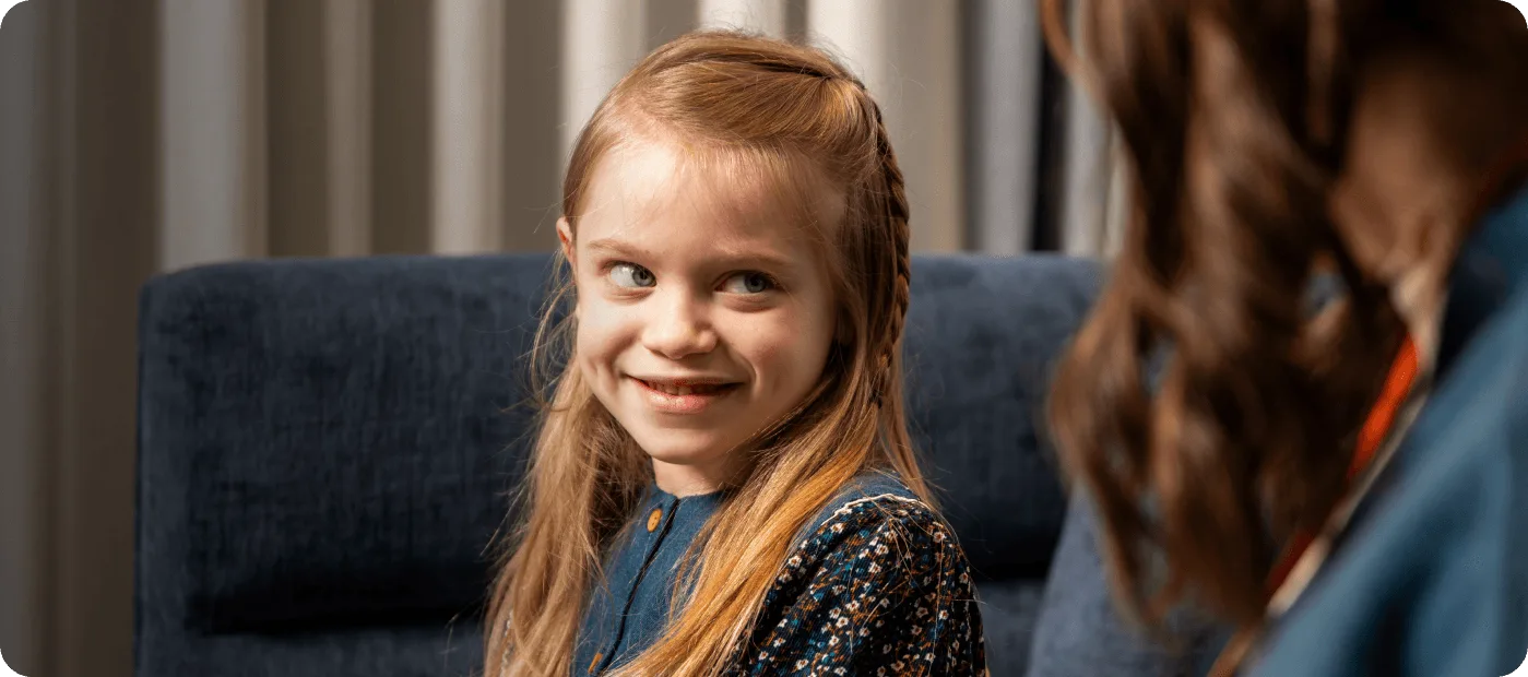 Young girl with long blonde hair smiling and looking at a person with brown hair.