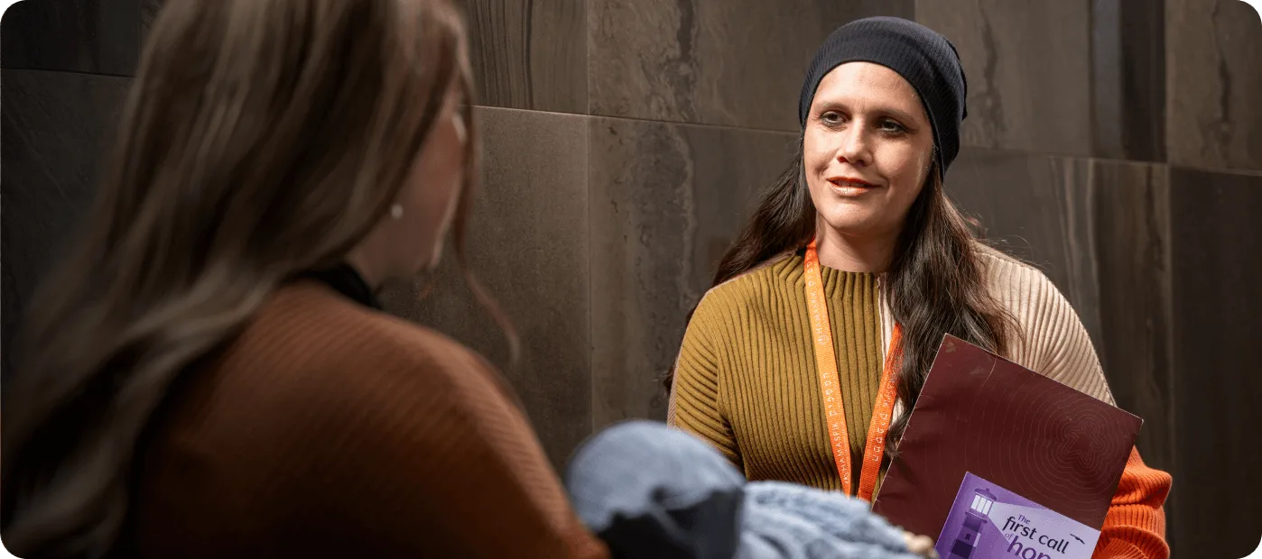 Smiling woman in a beanie speaking to another person holding a baby wrapped in a blue blanket.