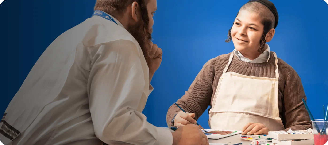 Boy in a white apron painting while an adult watches and talks with him against a blue background.