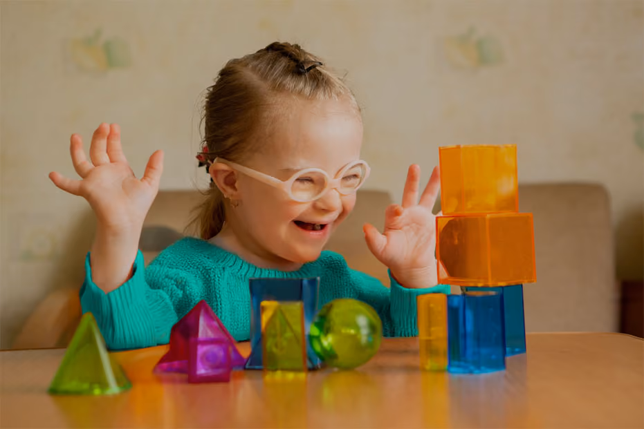 Smiling child with glasses playing with colorful translucent geometric blocks on a table.
