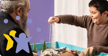 Smiling boy sprinkling sand on a play table with trees and animal toys while an older man watches.