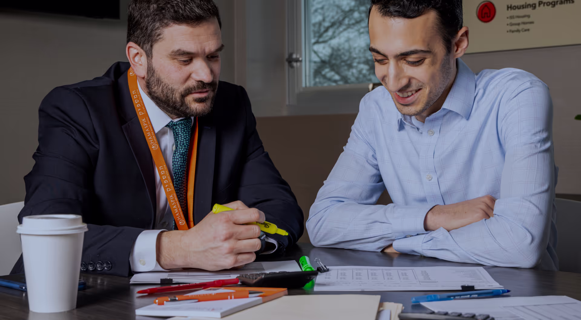 Two men reviewing documents at a desk, one highlighting text, the other smiling and watching.