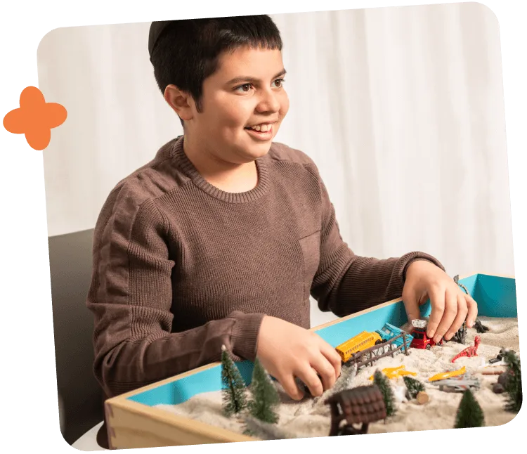 Smiling boy playing with a sand tray containing miniature trees, vehicles, and buildings.