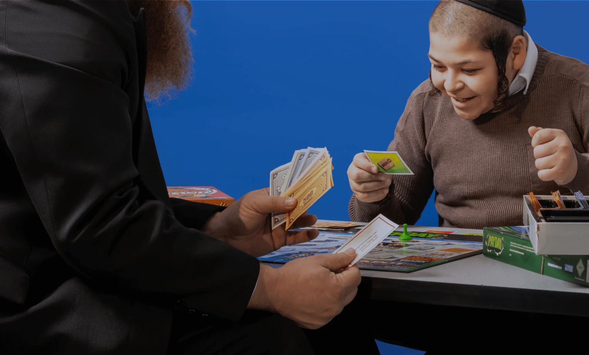 Two people playing a board game with money and cards, one person smiles excitedly.