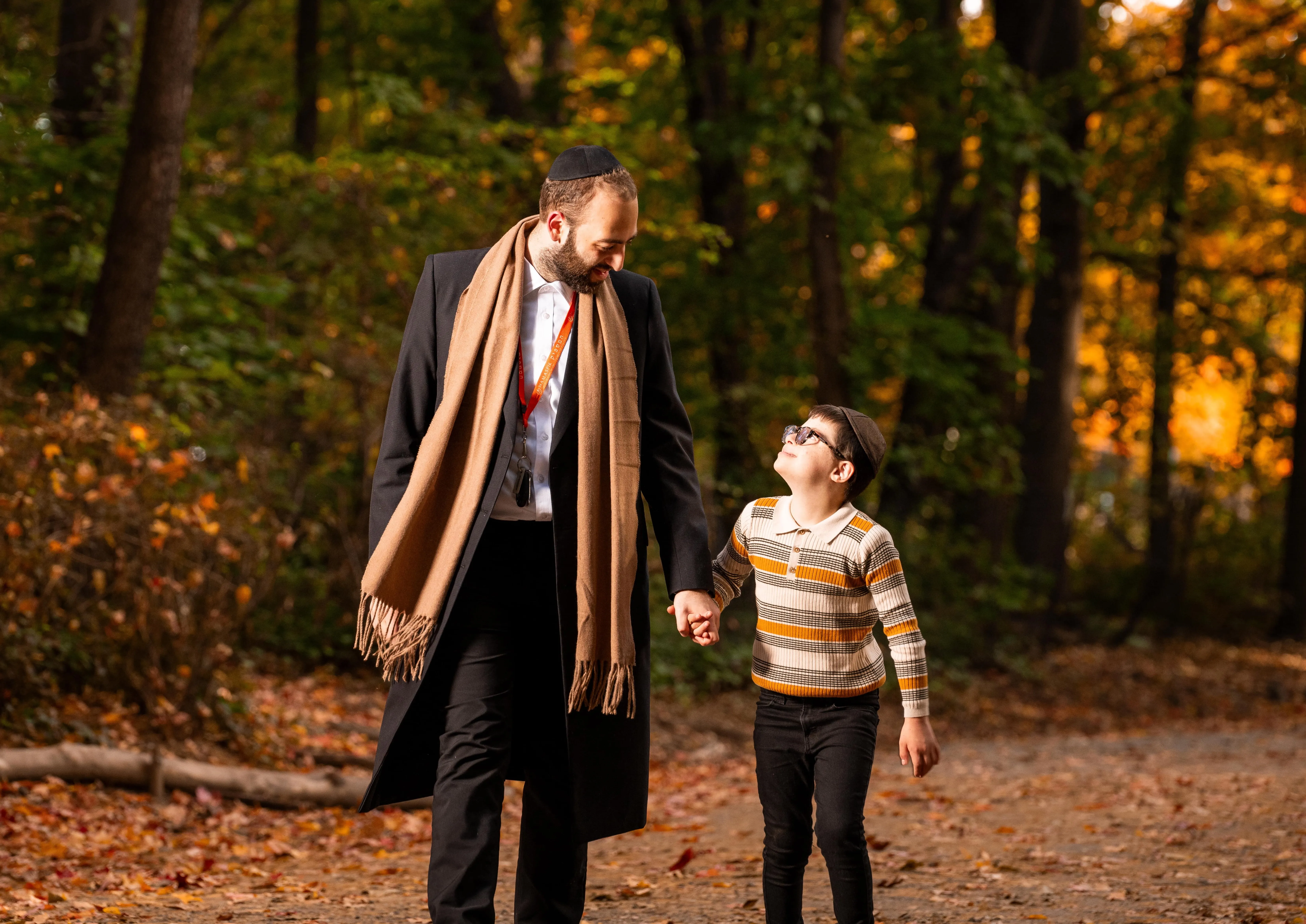 Man in coat and scarf holding hands with a boy wearing glasses and a striped sweater on a forest path.