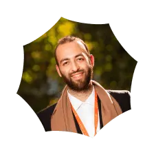 Smiling bearded man wearing a dark blazer and orange scarf outdoors with blurred greenery background.