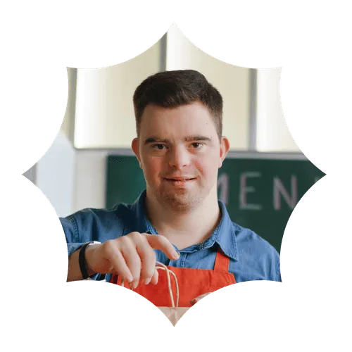 Smiling man with Down syndrome wearing an orange apron holding a paper bag in a kitchen.