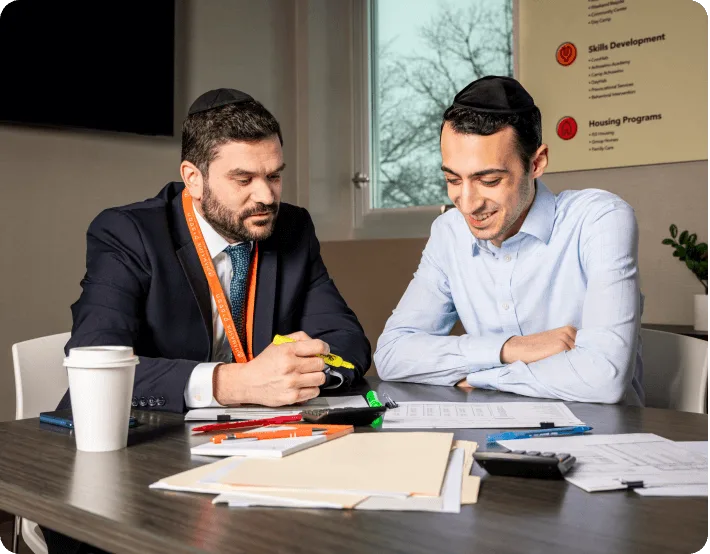 Two men wearing kippahs sit at a desk reviewing documents with pens and a coffee cup nearby.