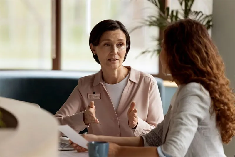 Woman in light pink shirt explaining or discussing documents with another woman in an office setting.