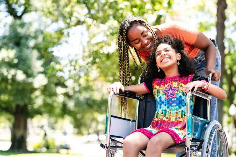 Smiling woman with long braids leans over young girl in colorful dress sitting in a wheelchair outdoors.