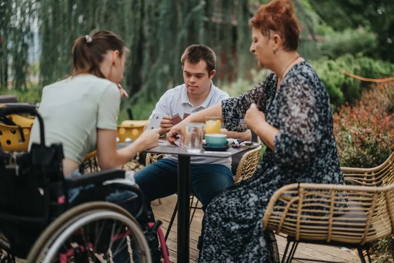 Three people, including a woman in a wheelchair, sitting outdoors playing cards at a table.