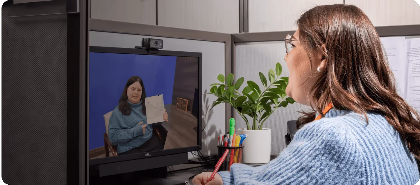 Woman in blue sweater video calls another woman holding a document in an office cubicle.