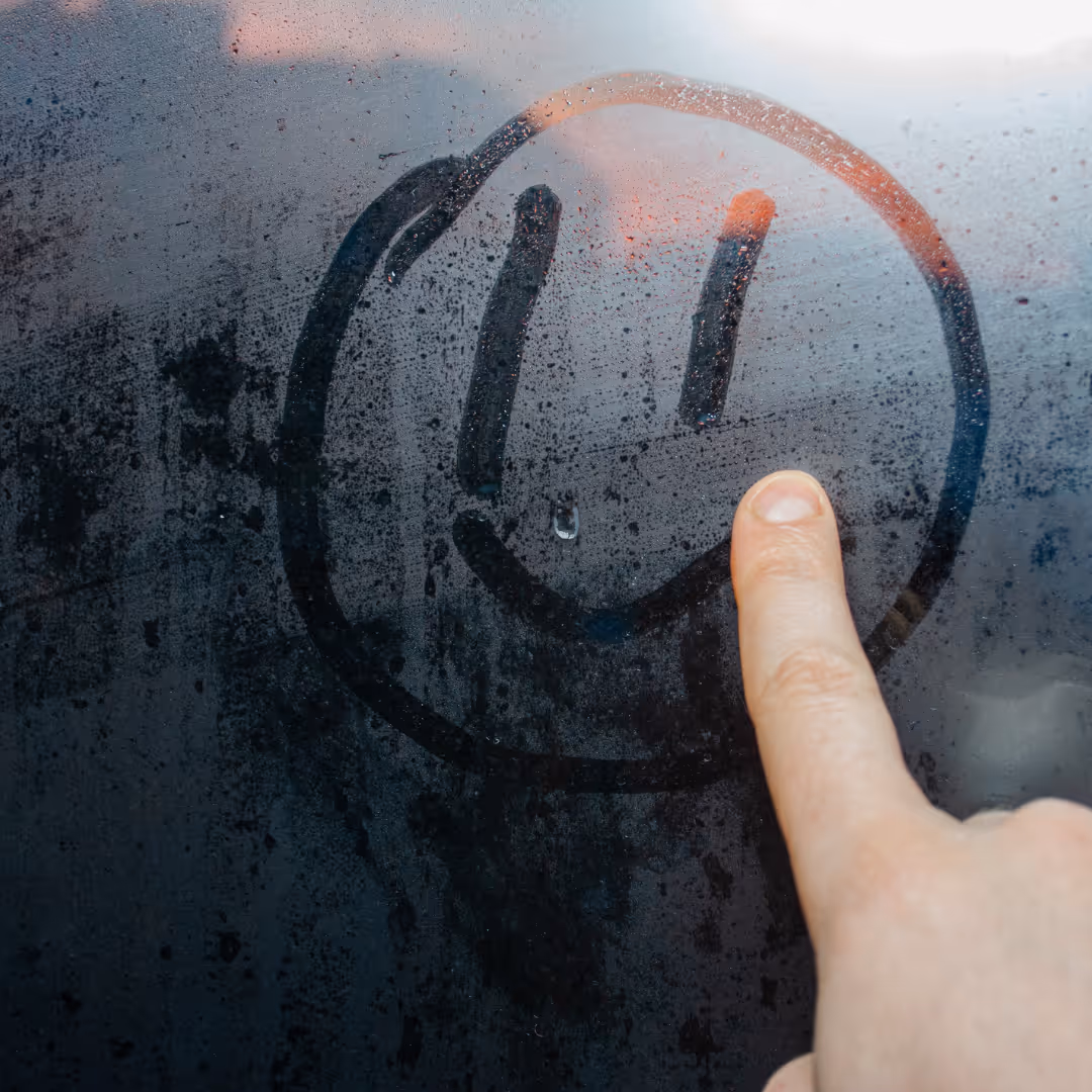 Finger drawing a smiley face on a foggy window with water droplets.