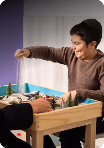 Boy smiling and sprinkling sand on a small wooden play table with toy trees and animals.