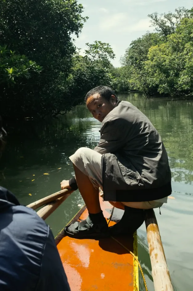 Person in a jacket and shorts sitting on the edge of a wooden boat on a river surrounded by green trees.