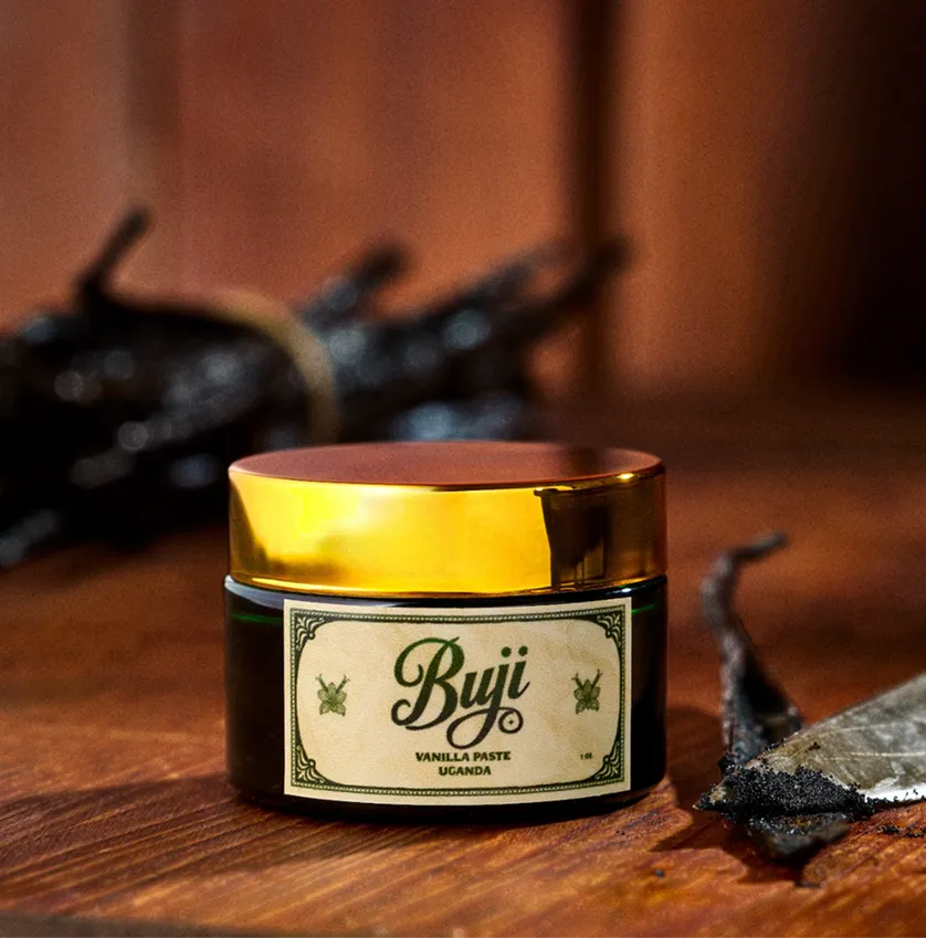 Jar of Byji vanilla paste from Uganda with golden lid on wooden surface with vanilla pods nearby.
