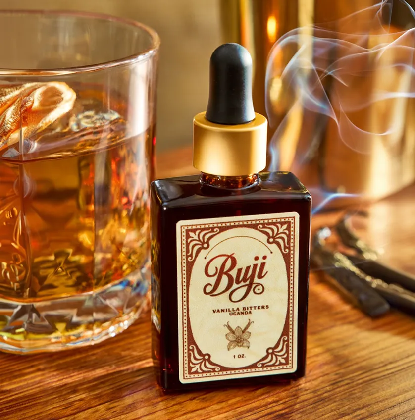Bottle of Buji Vanilla Bitters with dropper next to glass of whiskey and vanilla pods on wood table.