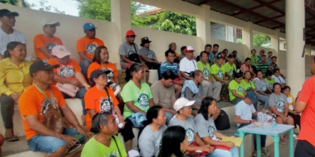 Group of people sitting on bleachers, some wearing orange, green, and gray shirts under a covered area.