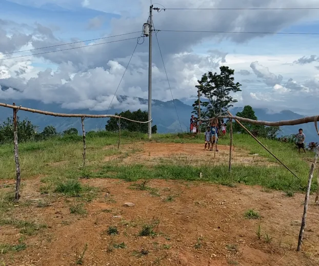 Children standing on a makeshift dirt volleyball court with wooden posts and natural scenery behind.