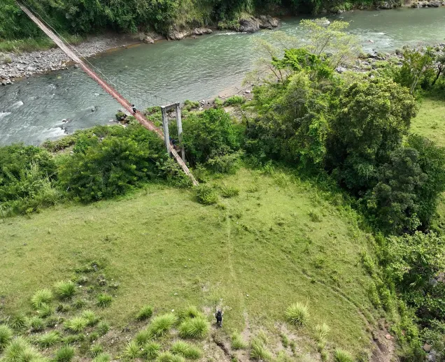 Person crossing a wooden suspension bridge over a river in a lush green landscape.