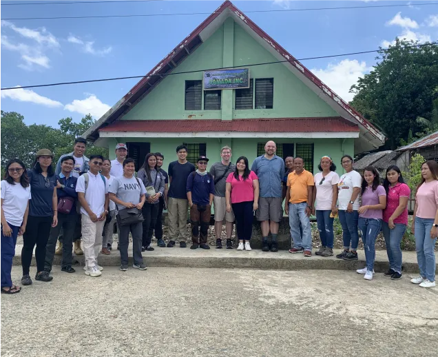 Group of 20 people standing in front of a green house with a red roof under a blue sky.