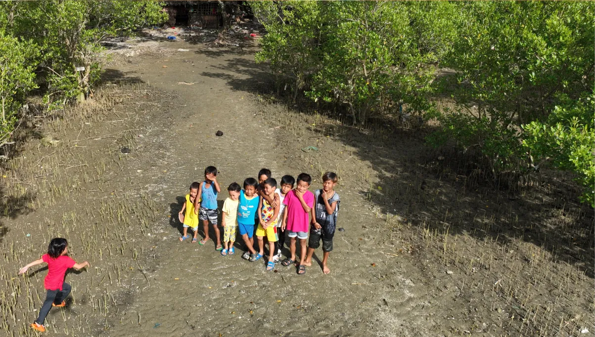 Group of children standing on muddy ground near mangroves, with one child running toward them.