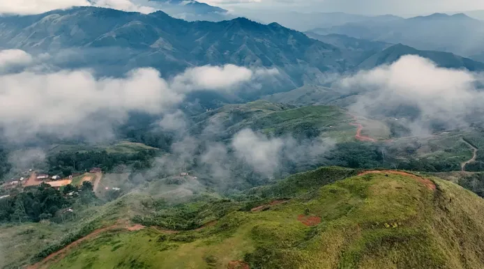 Mist-covered green hills and winding dirt roads under a cloudy sky in a mountainous landscape.
