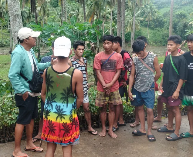 Group of young men standing and listening outdoors near palm trees and greenery in a tropical setting.