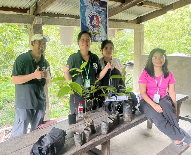 Four people pose with small tree seedlings in plastic bags on a wooden bench under a shelter.