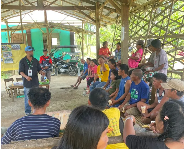 Man in blue cap speaking to seated group of people in an open wooden shelter during daytime.